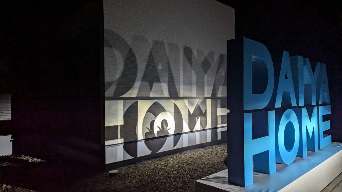 夜にライトアップされた「DAIYAHOME」の立体箱文字。背後の白い壁に文字の大きな影が映り、その影の前で人がピースサインをしている