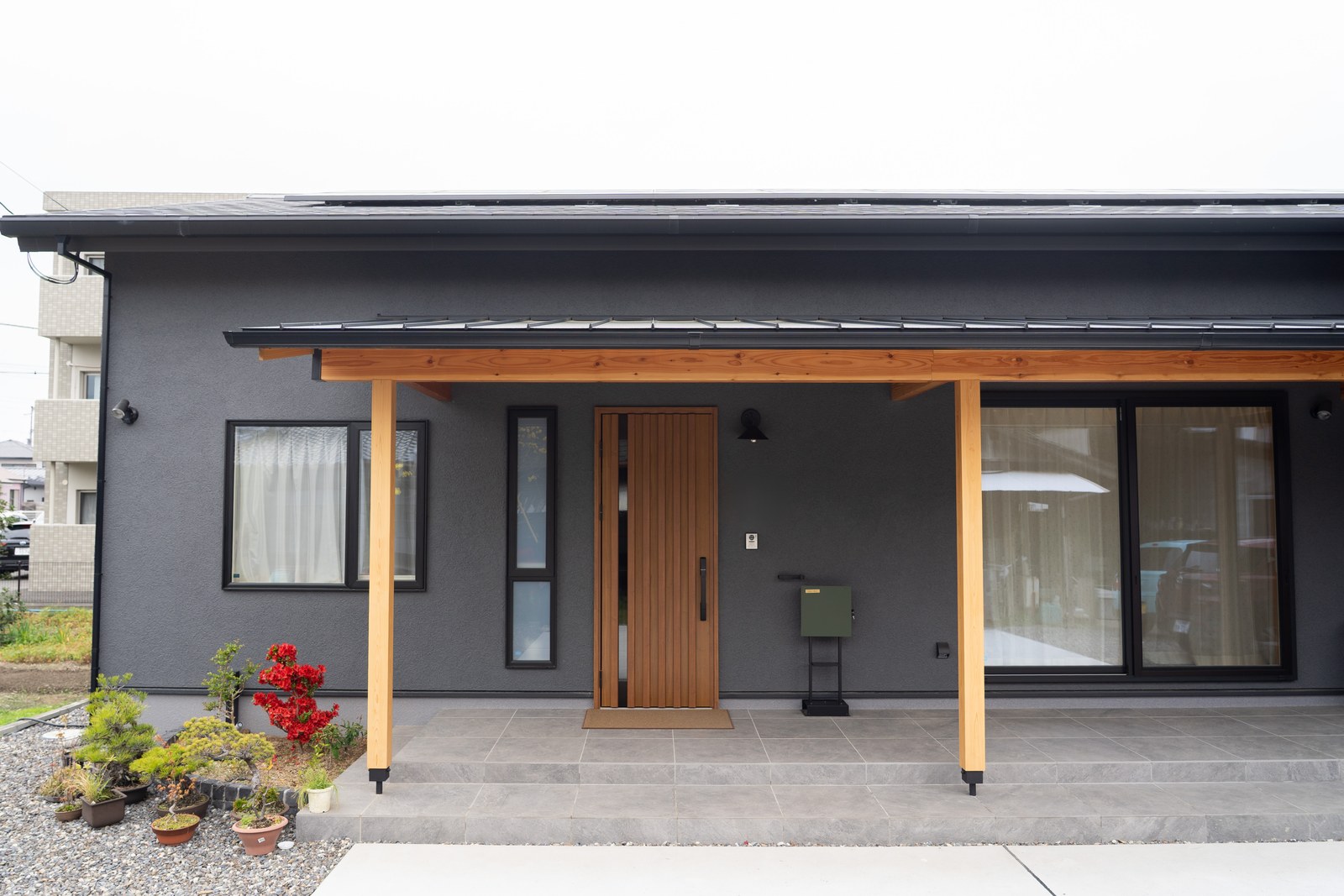 西条市の和風平屋の外観。チャコールグレーの塗り壁と木の柱、深い軒が続く玄関ポーチ