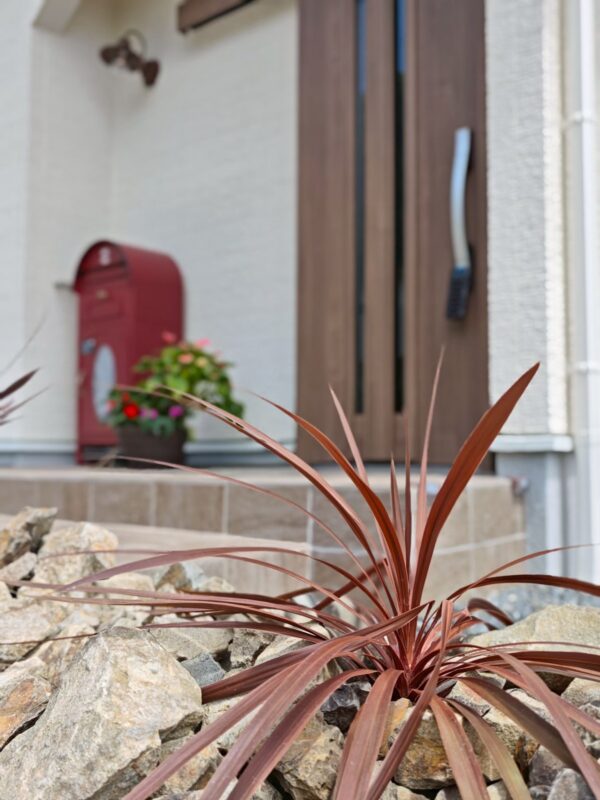 石積みの花壇に植えた赤葉の植栽越しに見る玄関ポーチ。赤いポストと寄せ植えが彩りを添える