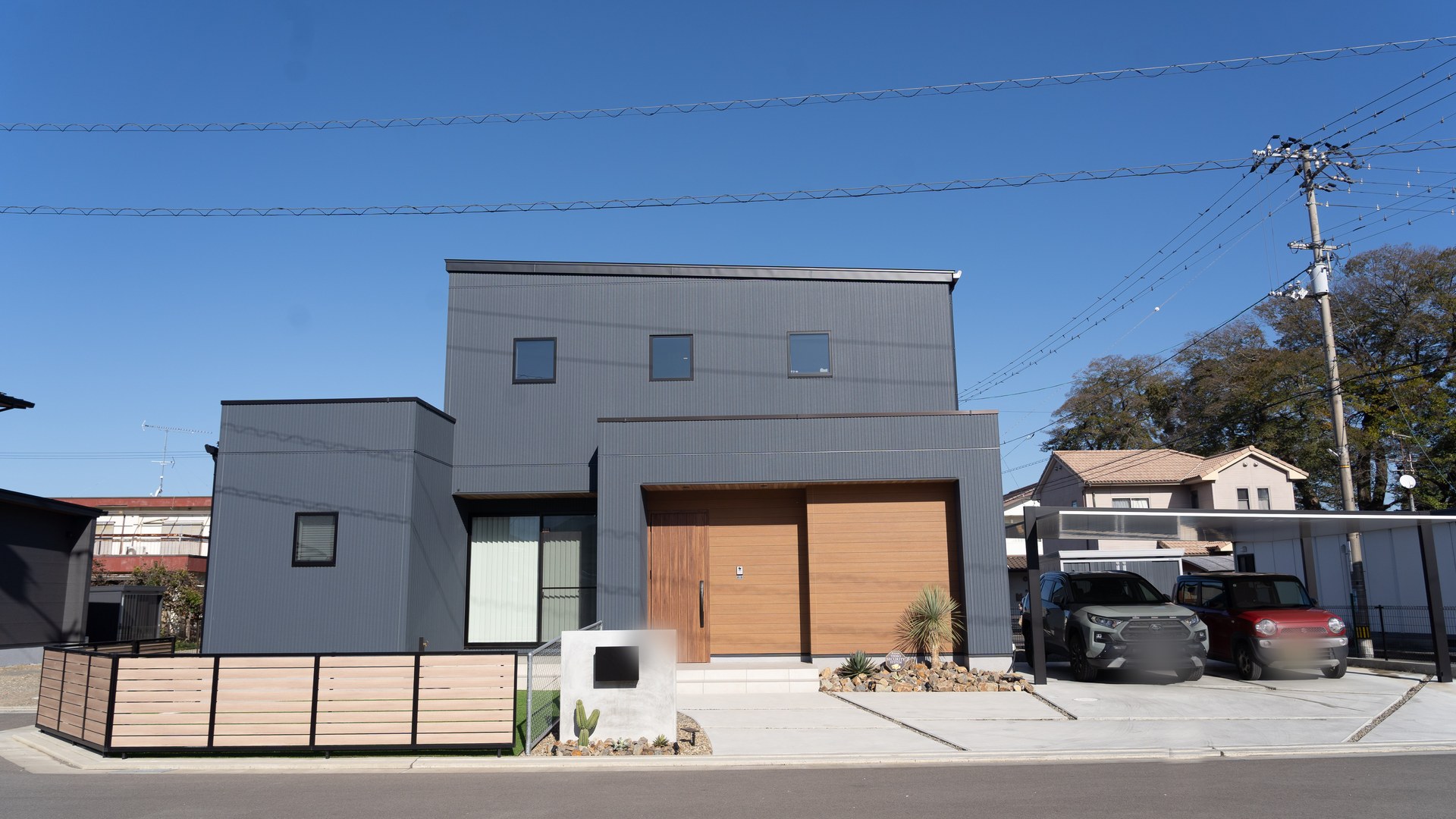 愛媛県新居浜市の2階建て注文住宅の外観。チャコールグレーの縦ラインの外壁に木目の玄関まわり、人工芝の庭とカーポート