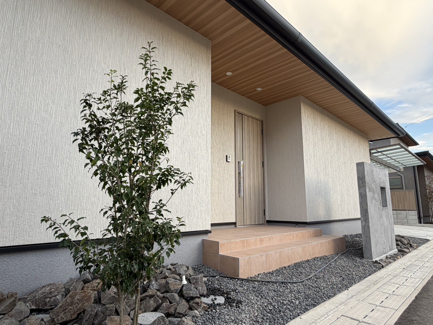白い外壁と木目の軒天、シンボルツリーと石の植栽が並ぶ玄関アプローチ